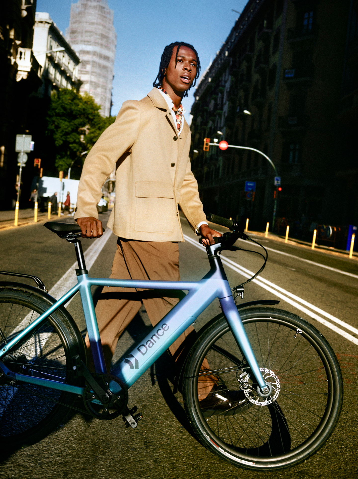 Man in a beige jacket standing confidently with a Dance e-bike on a wide city street in golden-hour light