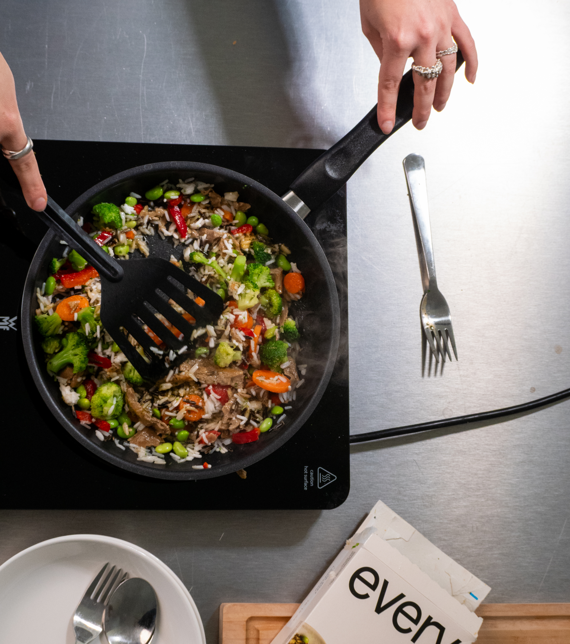 Overhead shot of hands stirring frozen food in a pan on an induction hob, with an Every Foods package next to it