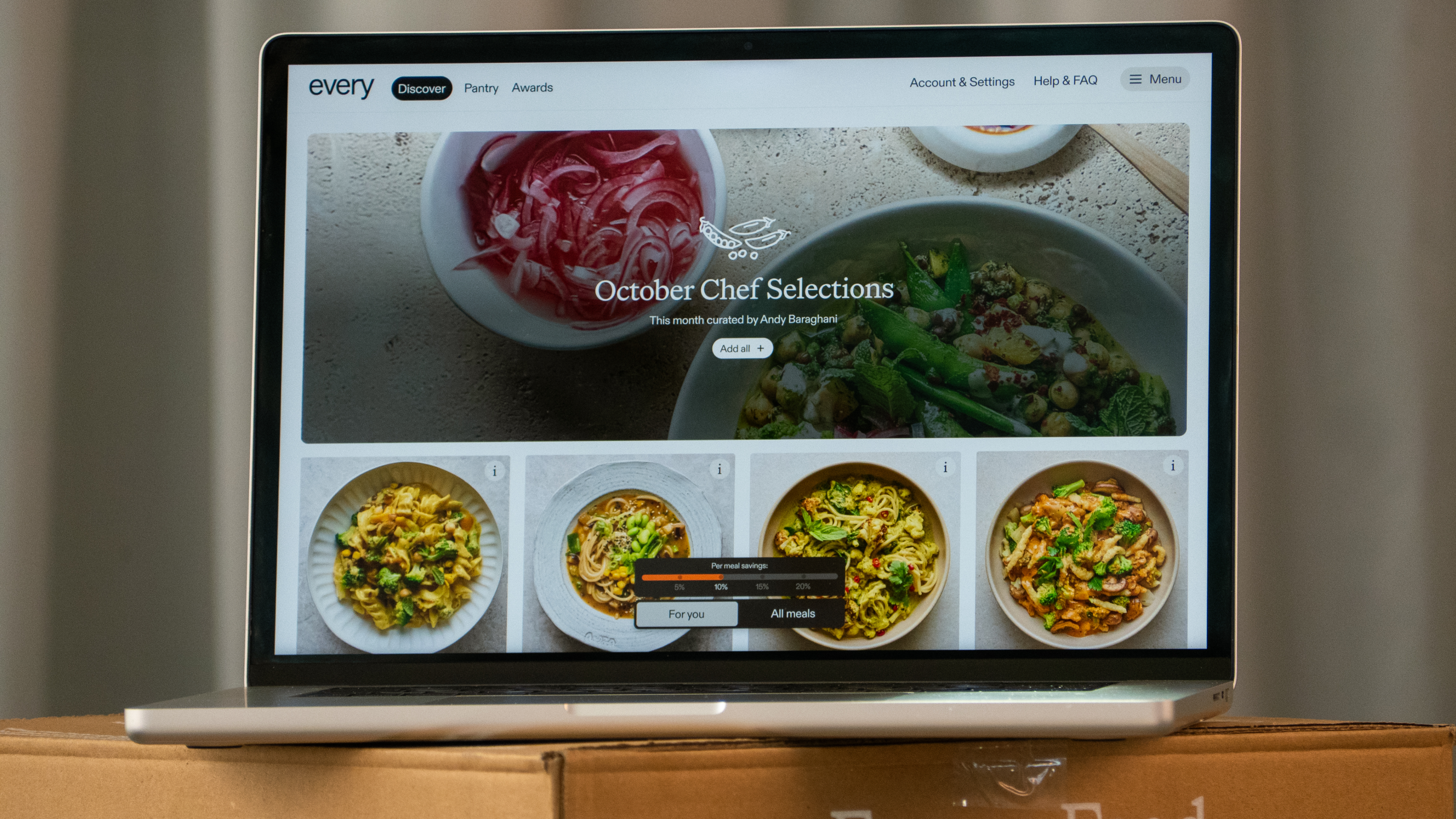 Macbook on an Every Foods delivery box showing the redesigned "Discover" homepage with an "October Chef Selections" hero banner and a personalized meal grid