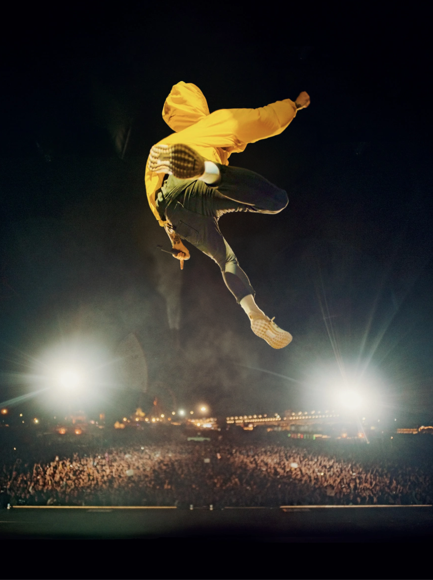 Casper in a yellow hoodie leaping high above a packed concert crowd, lit by bright stage lights.