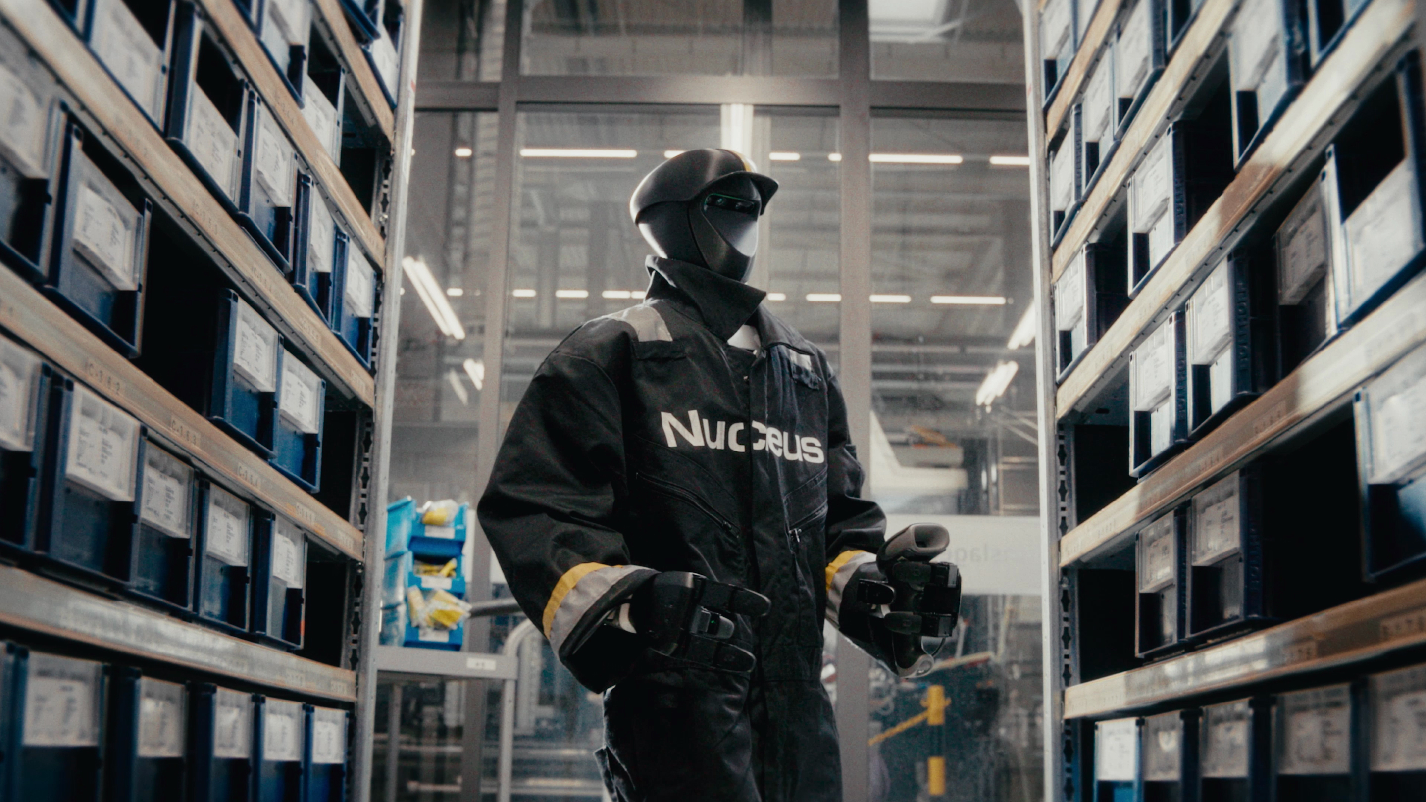 Humanoid robot in black Nucleus workwear and helmet standing between warehouse shelves stacked with blue storage bins