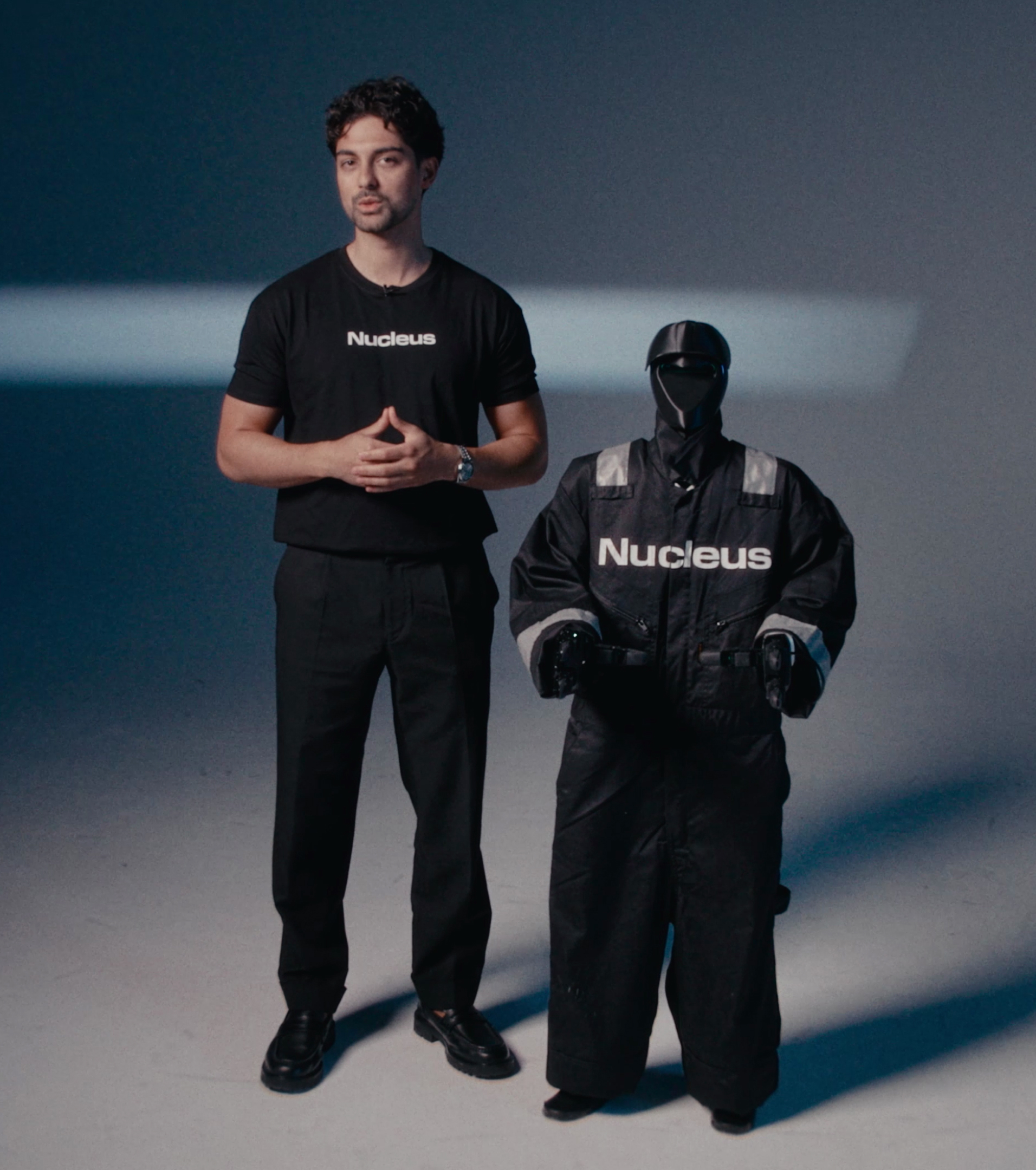 Man in a black Nucleus t-shirt standing beside a humanoid robot in black Nucleus coveralls in a softly lit studio