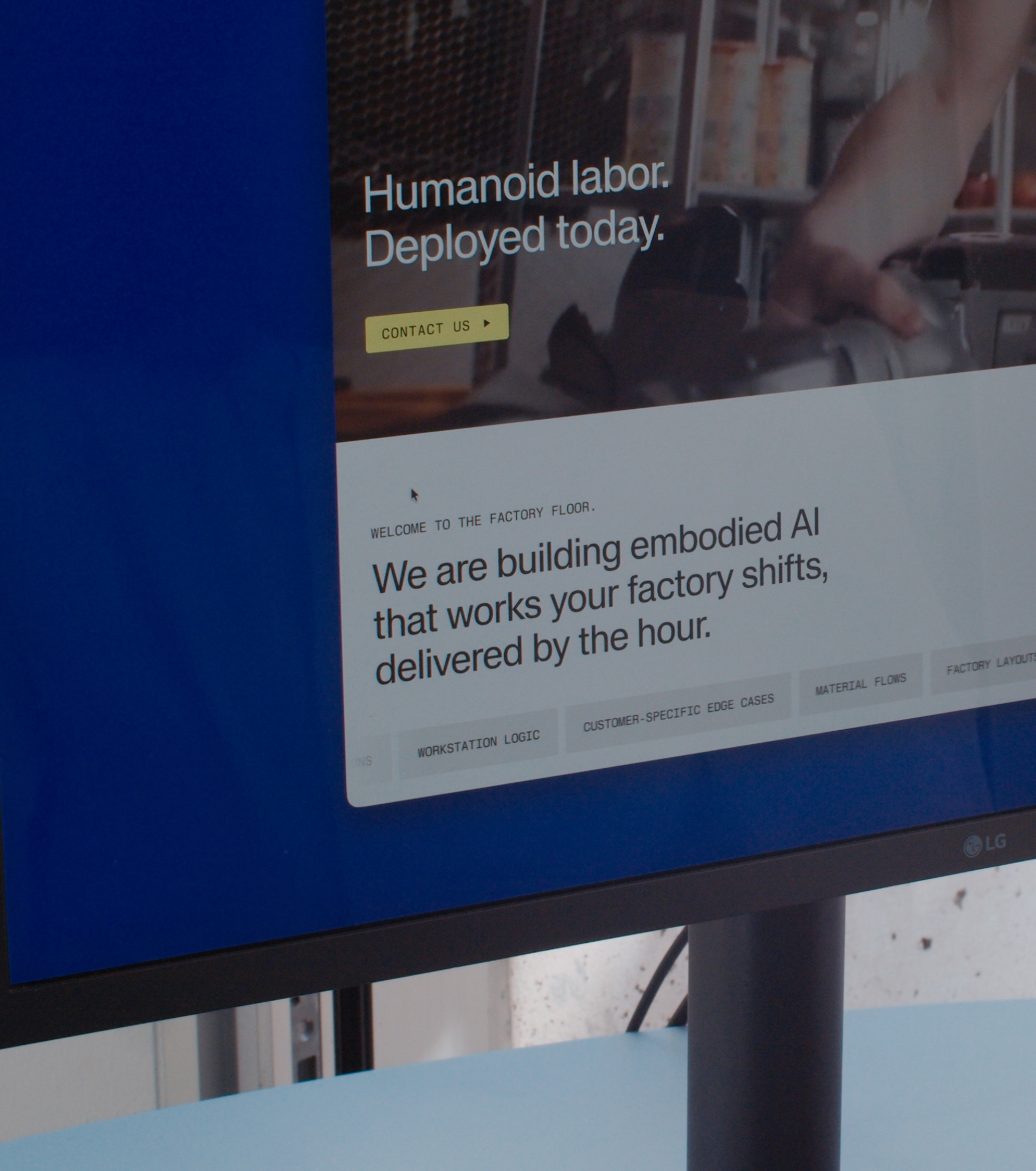 Monitor showing the Nucleus website with the headline "Humanoid labor. Deployed today." and "We are building embodied AI that works your factory shifts, delivered by the hour."