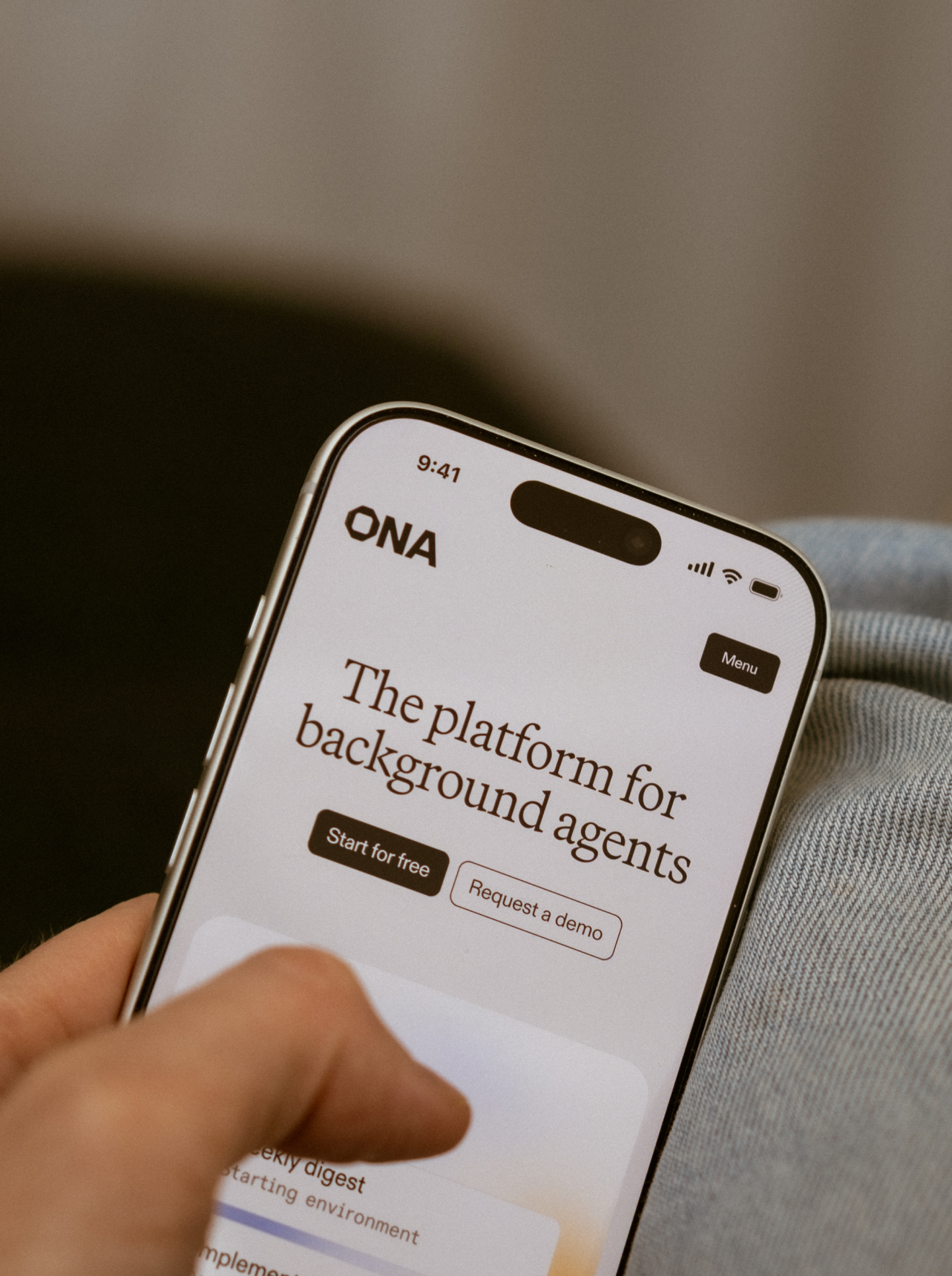 Hand holding an iPhone showing the Ona website homepage with the headline "The platform for background agents"