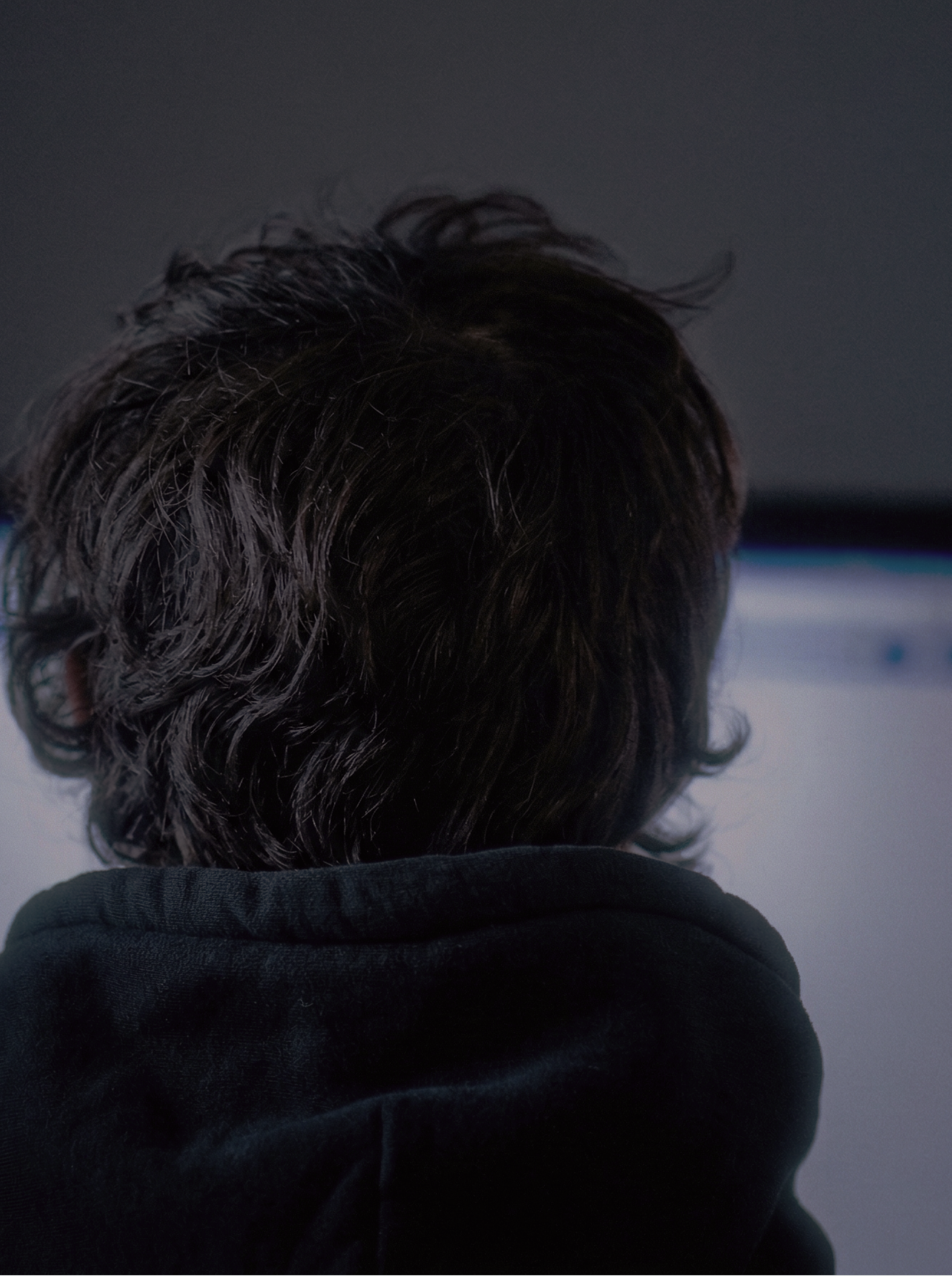 Close-up portrait from behind of a person with dark curly hair staring at a lit screen in a dark room