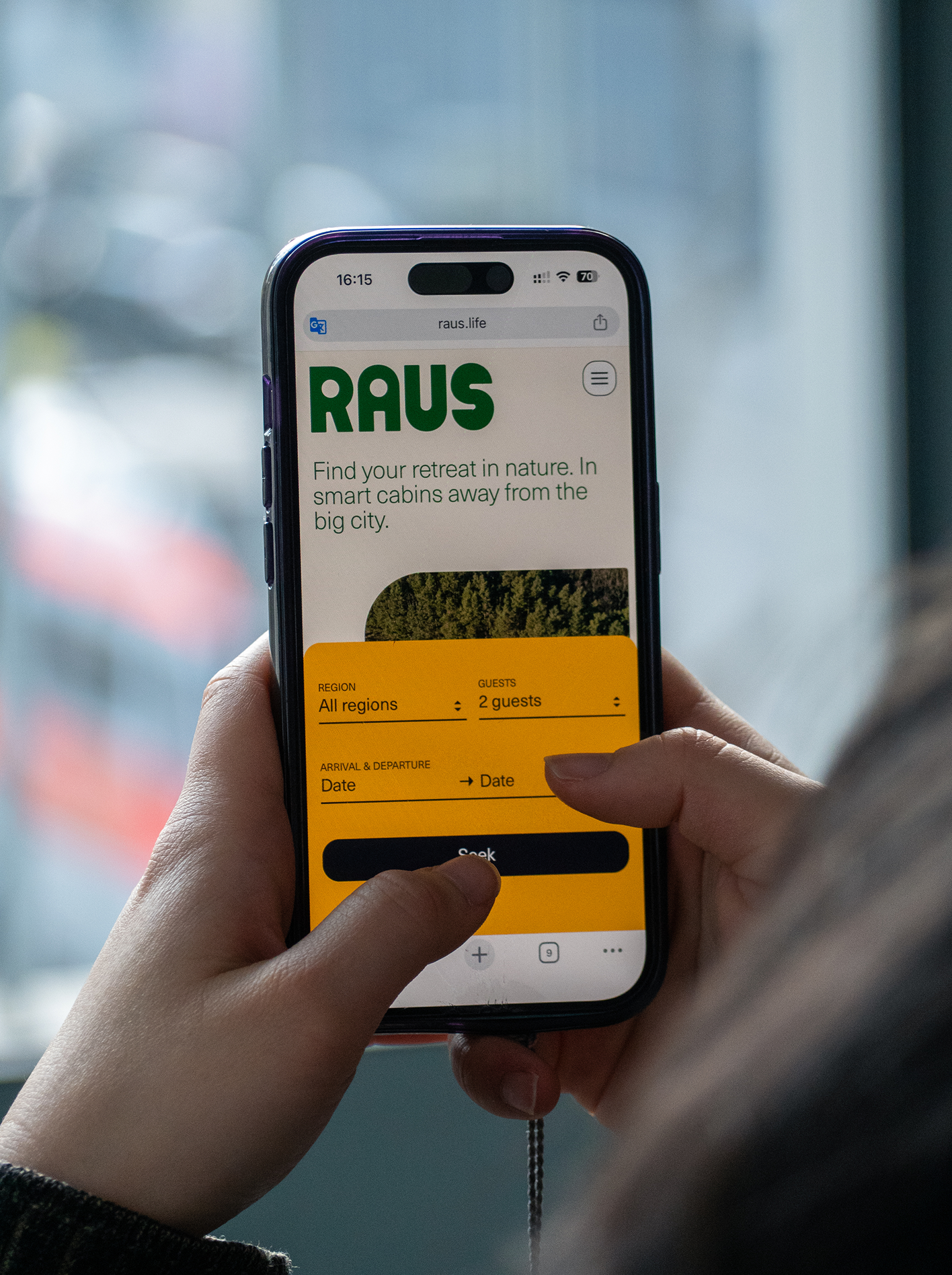 Hand holding an iPhone showing the Raus website with the search form for region, guests, arrival and departure date